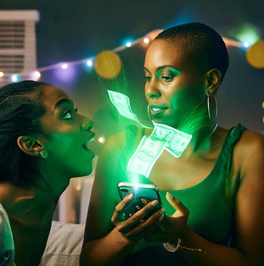 Two friends at a party reacting as one sprays money digitally from her phone, banknotes glowing green above the screen